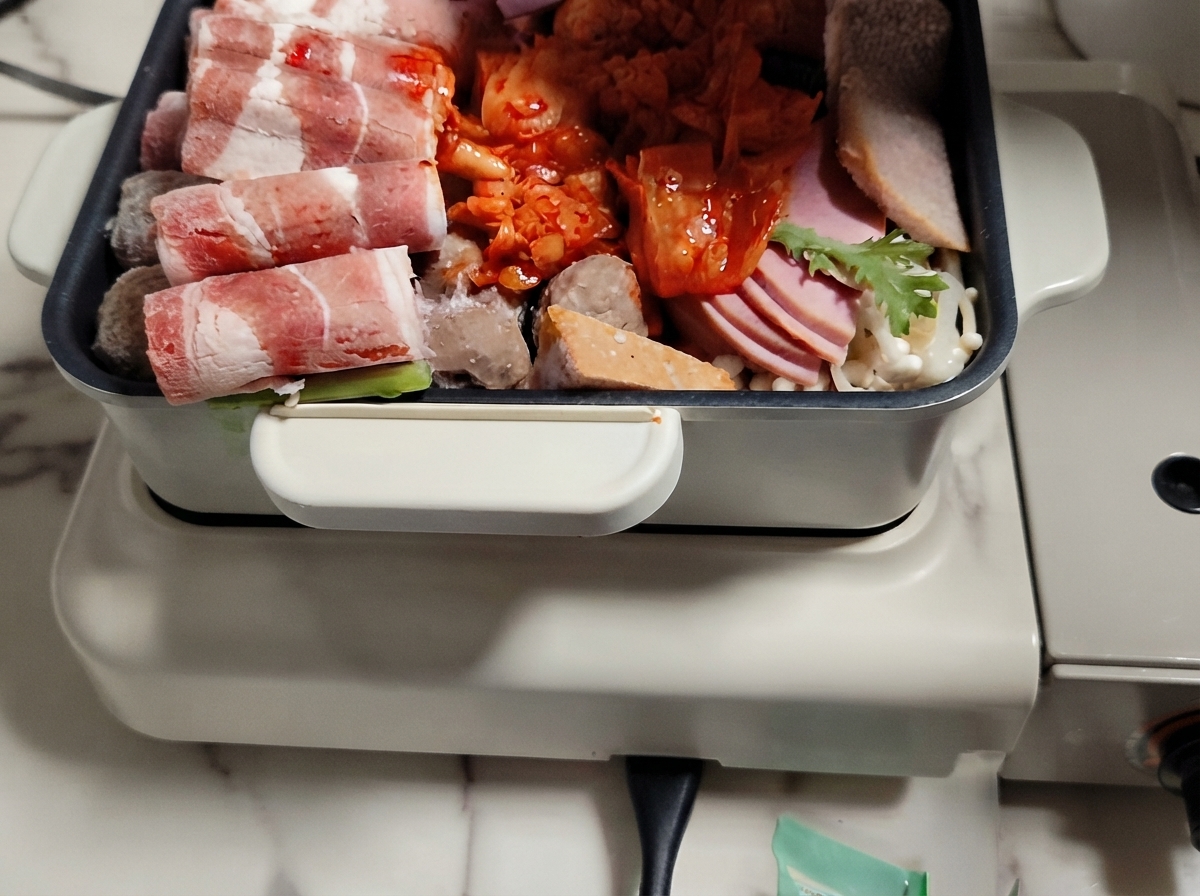 A hot pot filled with arranged layers of meat, vegetables, and rice cakes with broth being poured over them.