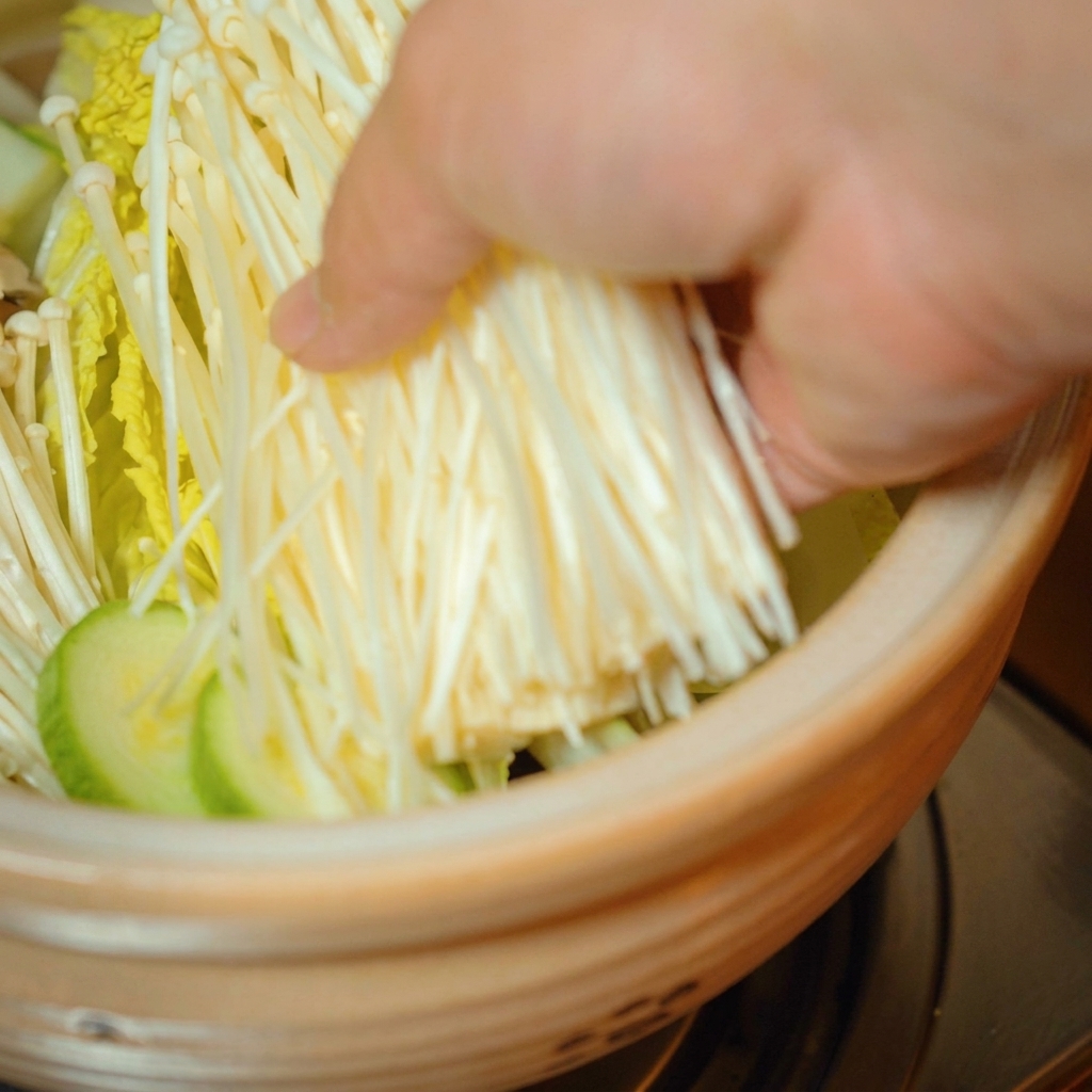 Fresh enoki mushrooms being placed on top of the vegetables in the pot.