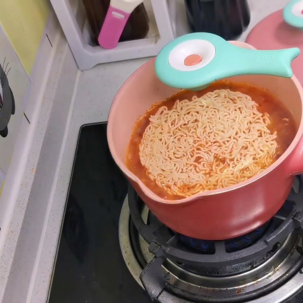 A package of instant noodles being dropped into a pink pot of boiling spicy red broth on a stove.