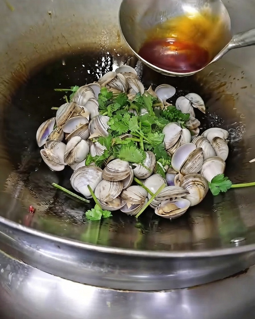 A ladle pouring chili oil and sesame oil over white clams and cilantro in a wok.