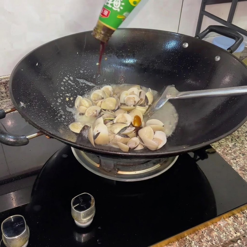 Oyster sauce flowing from a bottle into a wok filled with freshly opened white clams and cooking liquid.