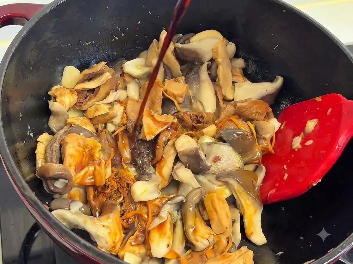 Light soy sauce being poured from above onto the mixture of mushrooms in the pot, with a red spatula nearby.