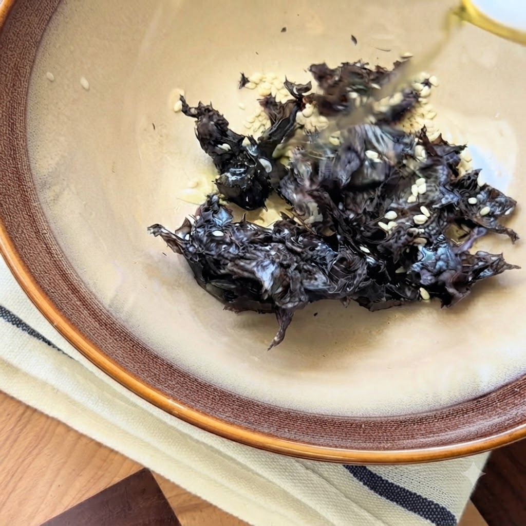 Sichuan pepper oil being poured over dried seaweed and white sesame seeds in a ceramic bowl.