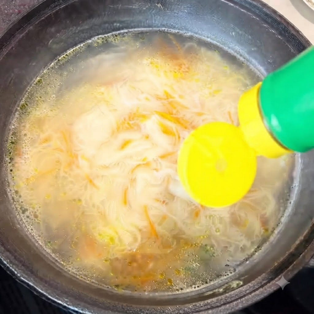 Oyster sauce being squeezed from a green-capped bottle into the cooking noodle soup.