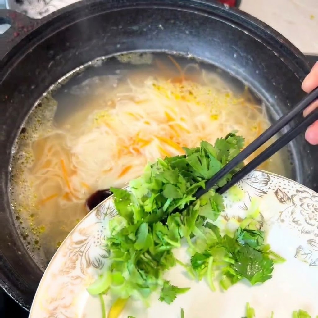 Chopped cilantro and scallions being dropped from a plate into the pot of noodles using chopsticks.