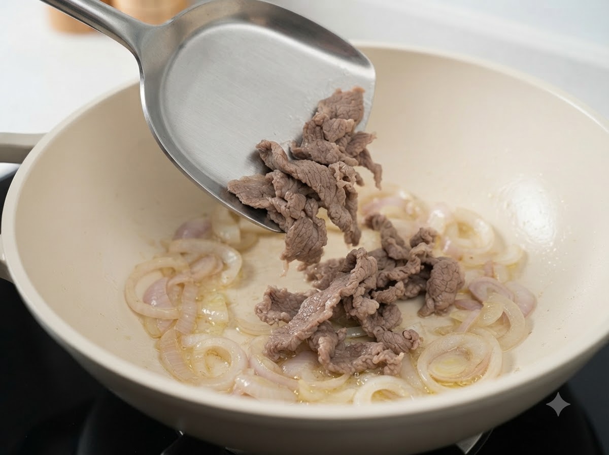 Blanched beef slices being dropped from a spatula into a white pan with sautéed sliced onions.