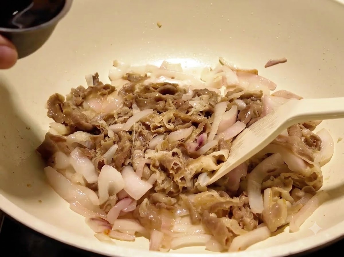 Dark liquid sauce being poured from a small metal bowl over beef and onions inside a white cooking pan.