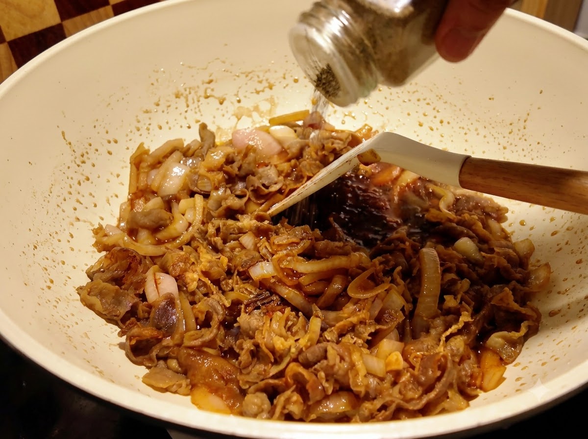 Black pepper being shaken from a glass container onto beef and onions simmering in sauce inside a pan.