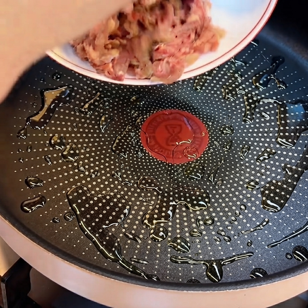 Raw ground beef being dropped from a plate into a frying pan with pools of cooking oil.