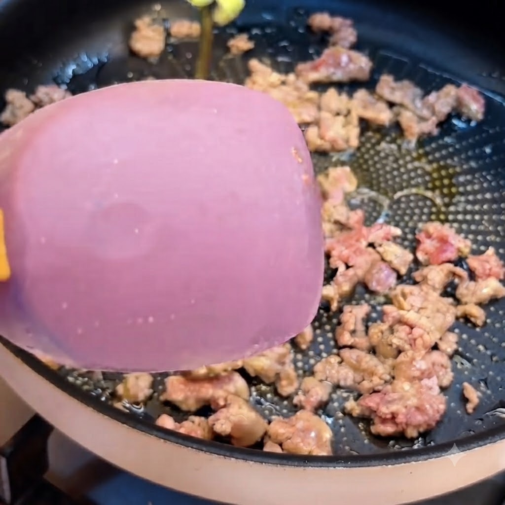 A pink spatula moving through ground beef being stir-fried in a dark frying pan.