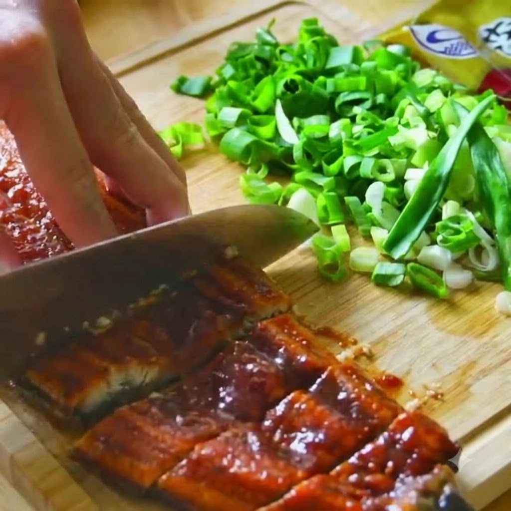 Grilled eel fillets being cut with a knife on a wooden cutting board near chopped green onions.