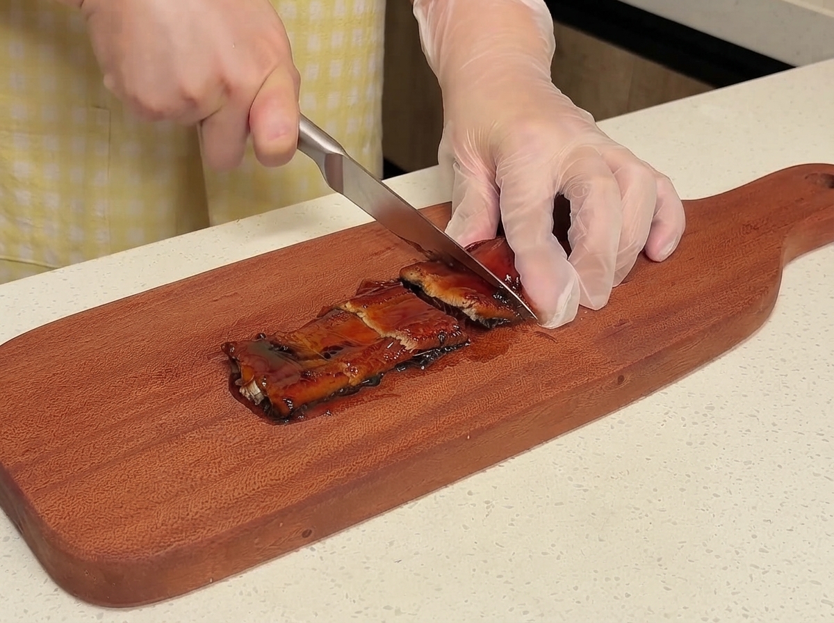 A person wearing gloves using a knife to slice pre-cooked eel on a wooden board.