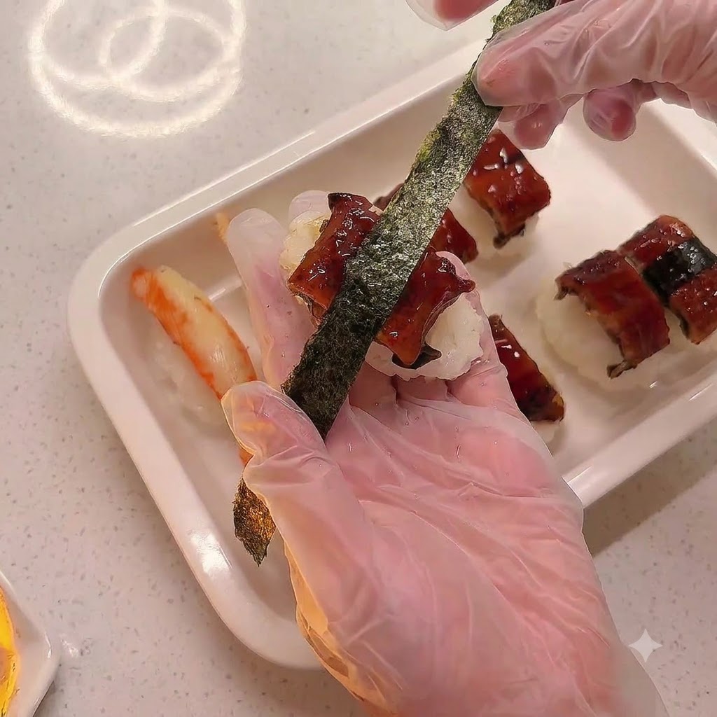 A gloved hand wrapping a strip of dried seaweed around an eel sushi piece placed on a white tray.