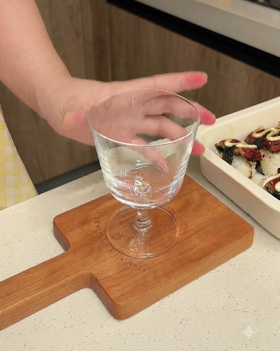 A clear stemmed glass sitting on a small wooden cutting board next to a tray of prepared sushi.