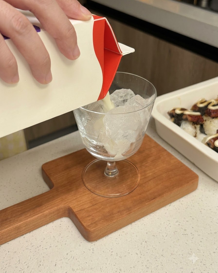 Milk being poured from a carton into a clear glass containing ice cubes.