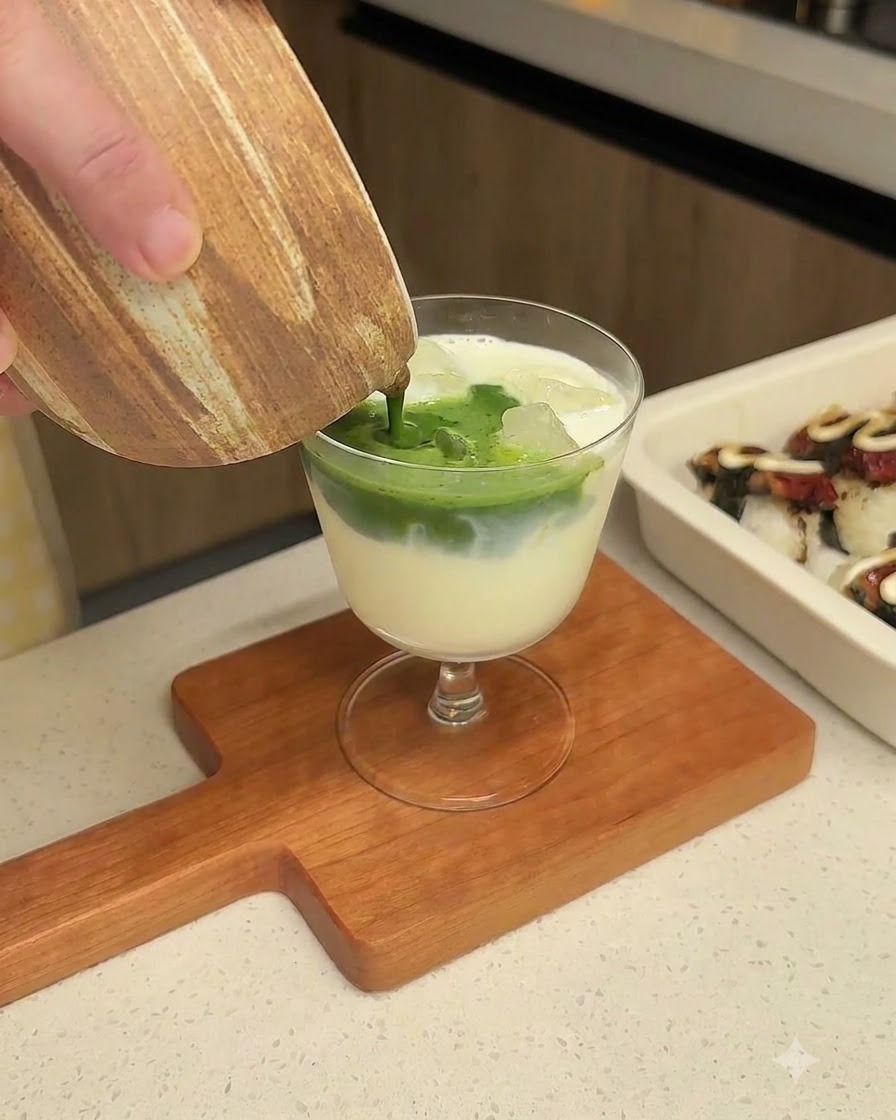 Dark green whisked matcha being poured from a ceramic pitcher into a glass filled with milk and ice.