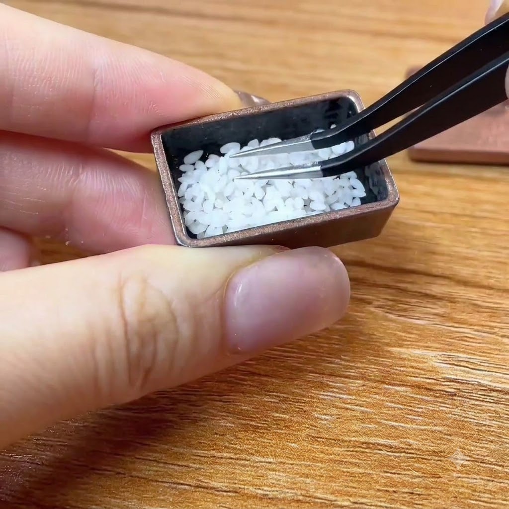 Tweezers placing tiny white rice grains into a miniature brown container.