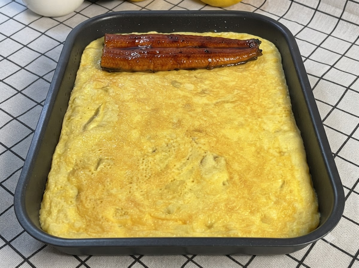 A piece of glazed cooked eel placed on top of a thin cooked egg layer in a rectangular pan.
