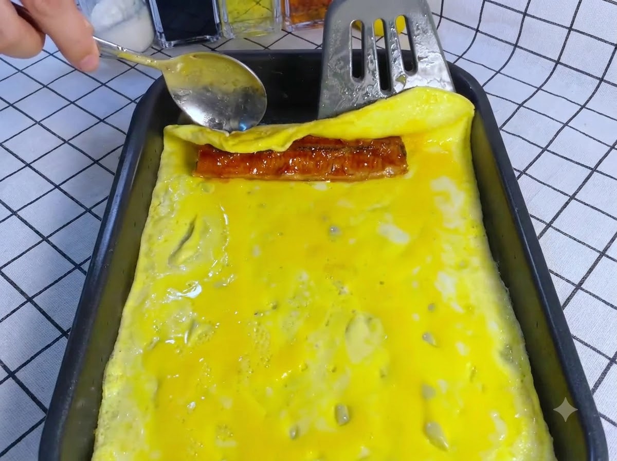 A spatula rolling the cooked egg layer over a piece of eel inside a rectangular pan.