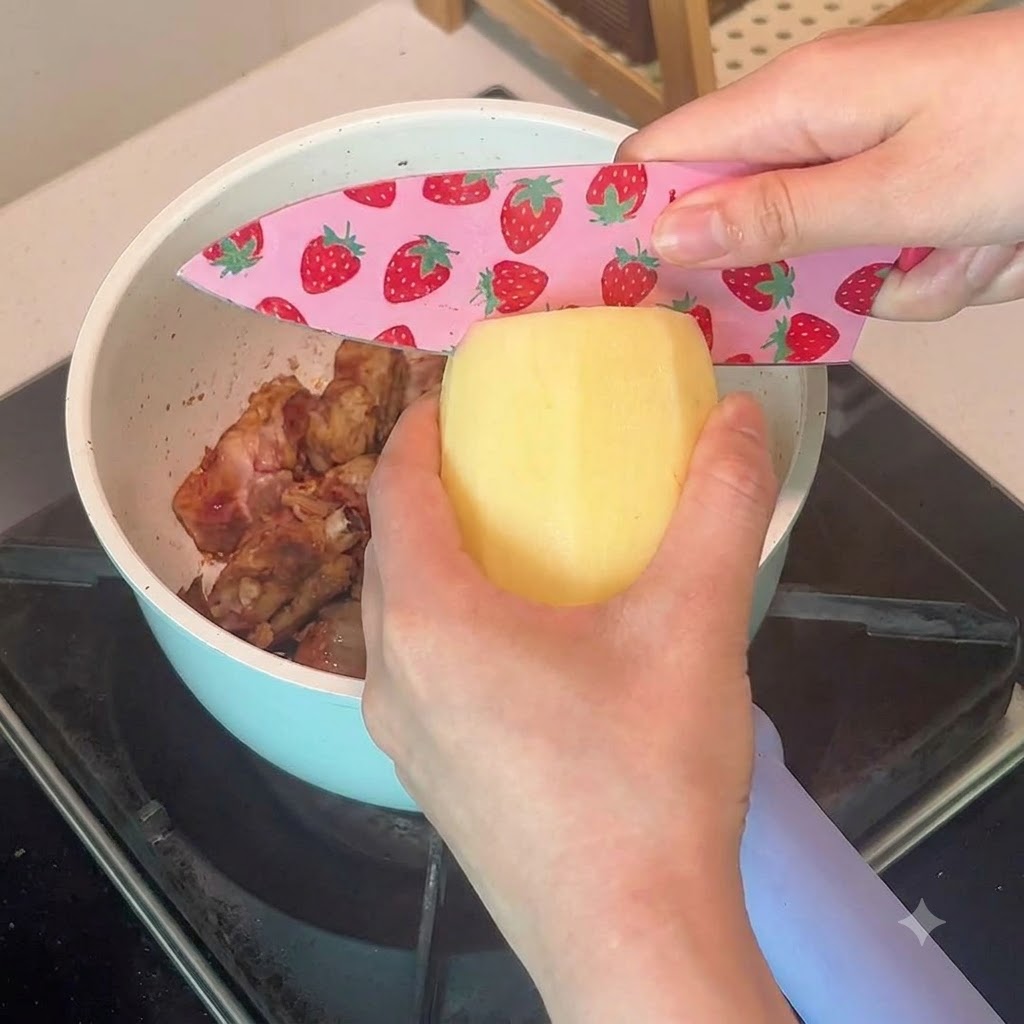 A peeled potato being sliced with a pink strawberry-patterned knife over a pot of pork ribs.