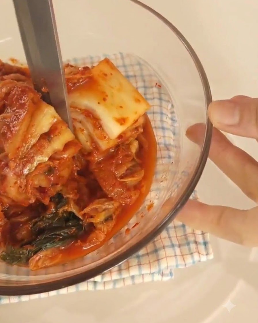 Kitchen scissors cutting pieces of kimchi inside a clear glass bowl lined with a checkered cloth.