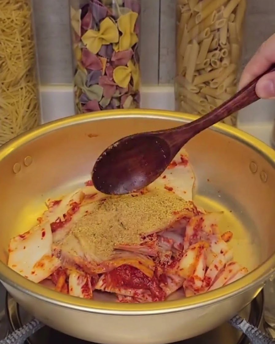 Chopped pieces of kimchi mixed with seasoning powder in a pot, being stirred with a dark wooden spoon while glass jars of dried pasta sit in the blurred background.