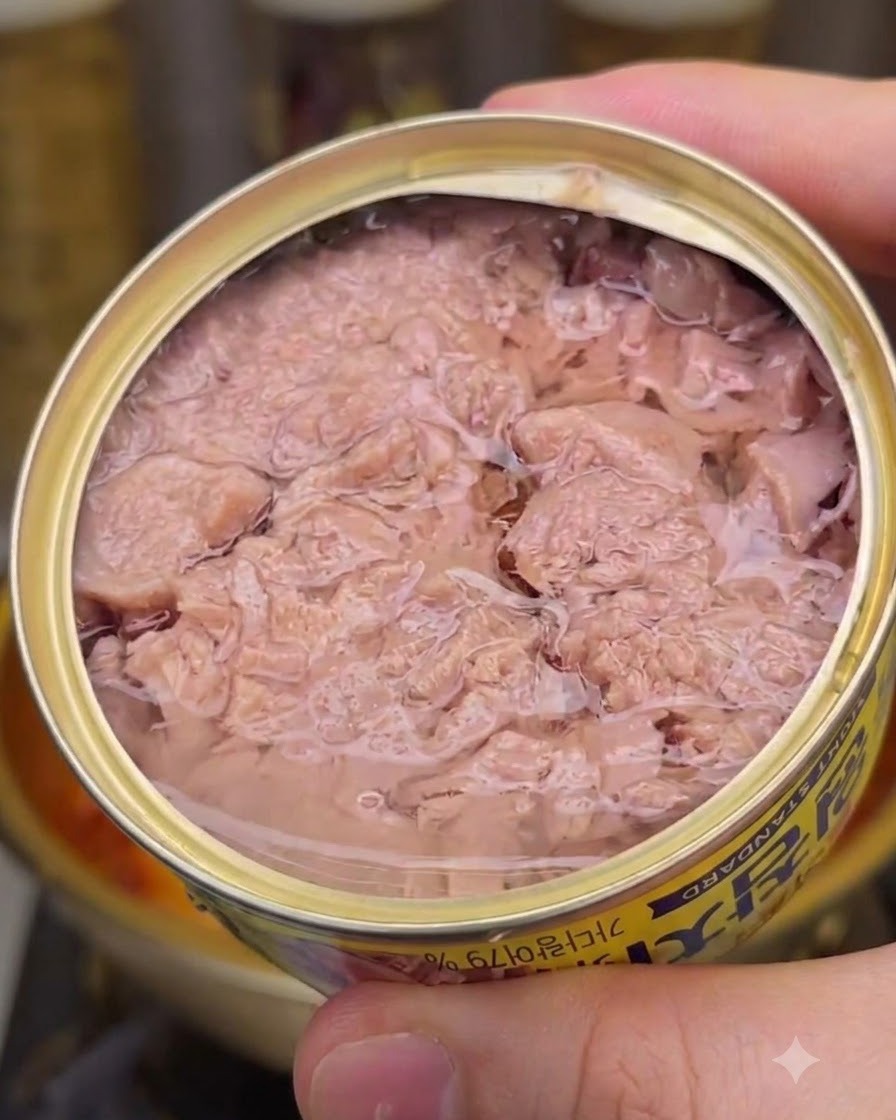 An open tin can filled with pink flaked tuna chunks and canning liquid, held up closely with the edge of a simmering pot visible underneath.