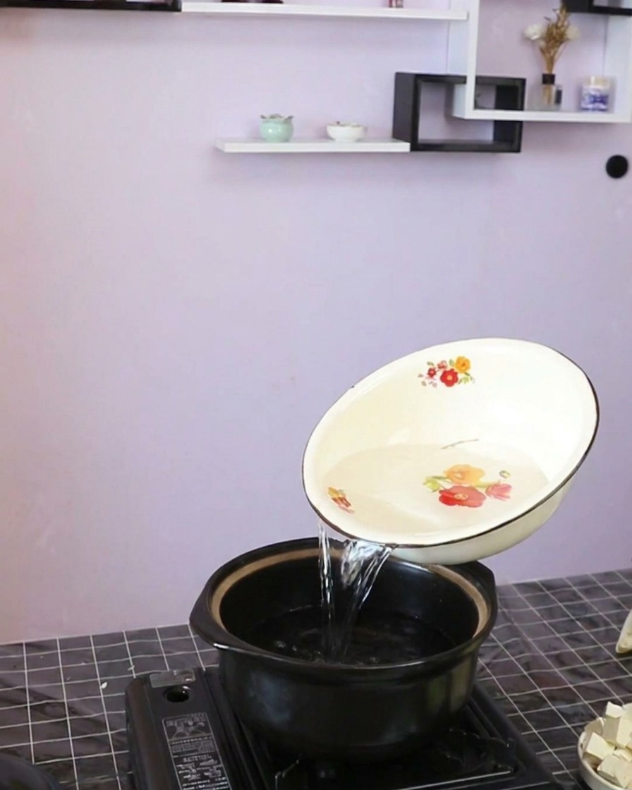 Clear water being poured from a decorated white bowl into a dark clay cooking pot.