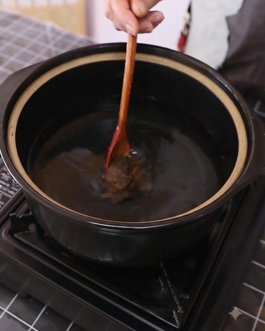 Soybean paste being stirred into water inside a dark cooking pot using a wooden spoon.