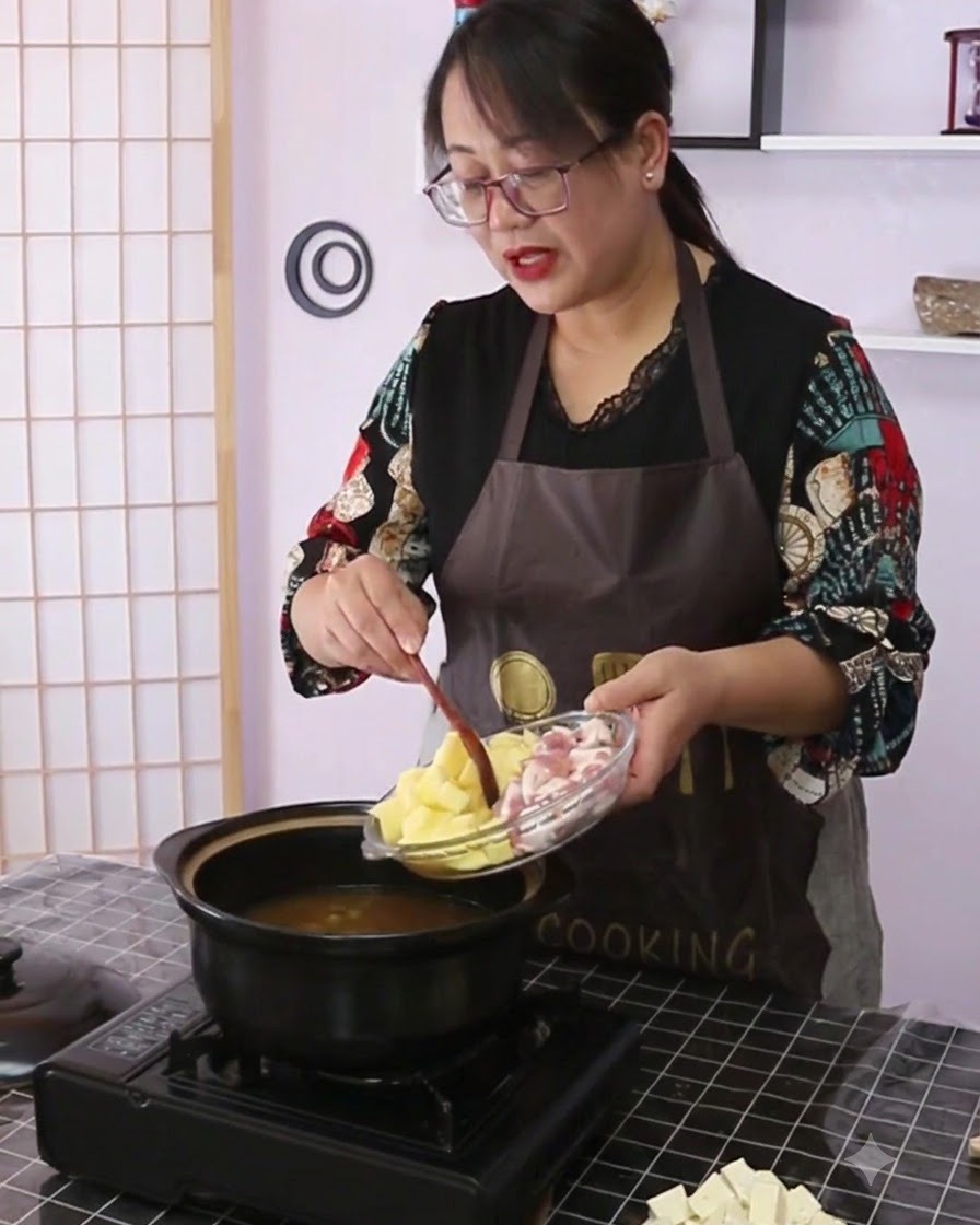 Potatoes and pork belly being poured from a glass plate into a black ceramic cooking pot filled with broth.
