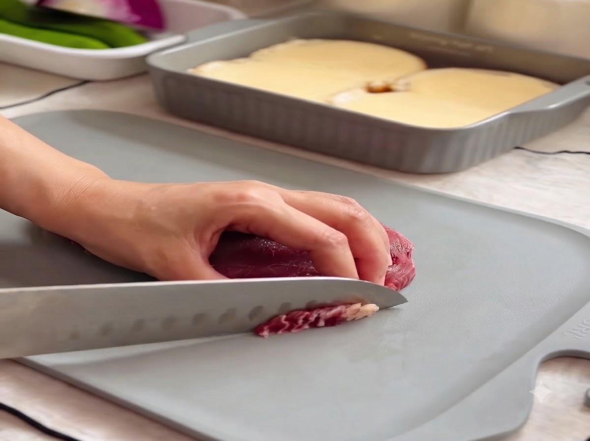 A hand holding a knife slicing raw ribeye steak into thin pieces on a grey cutting board.