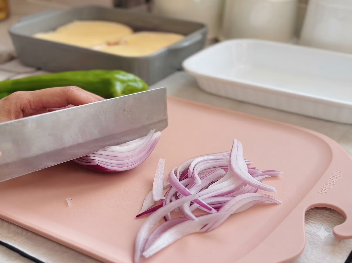 A kitchen knife slicing a red onion on a pink cutting board next to a green pepper.