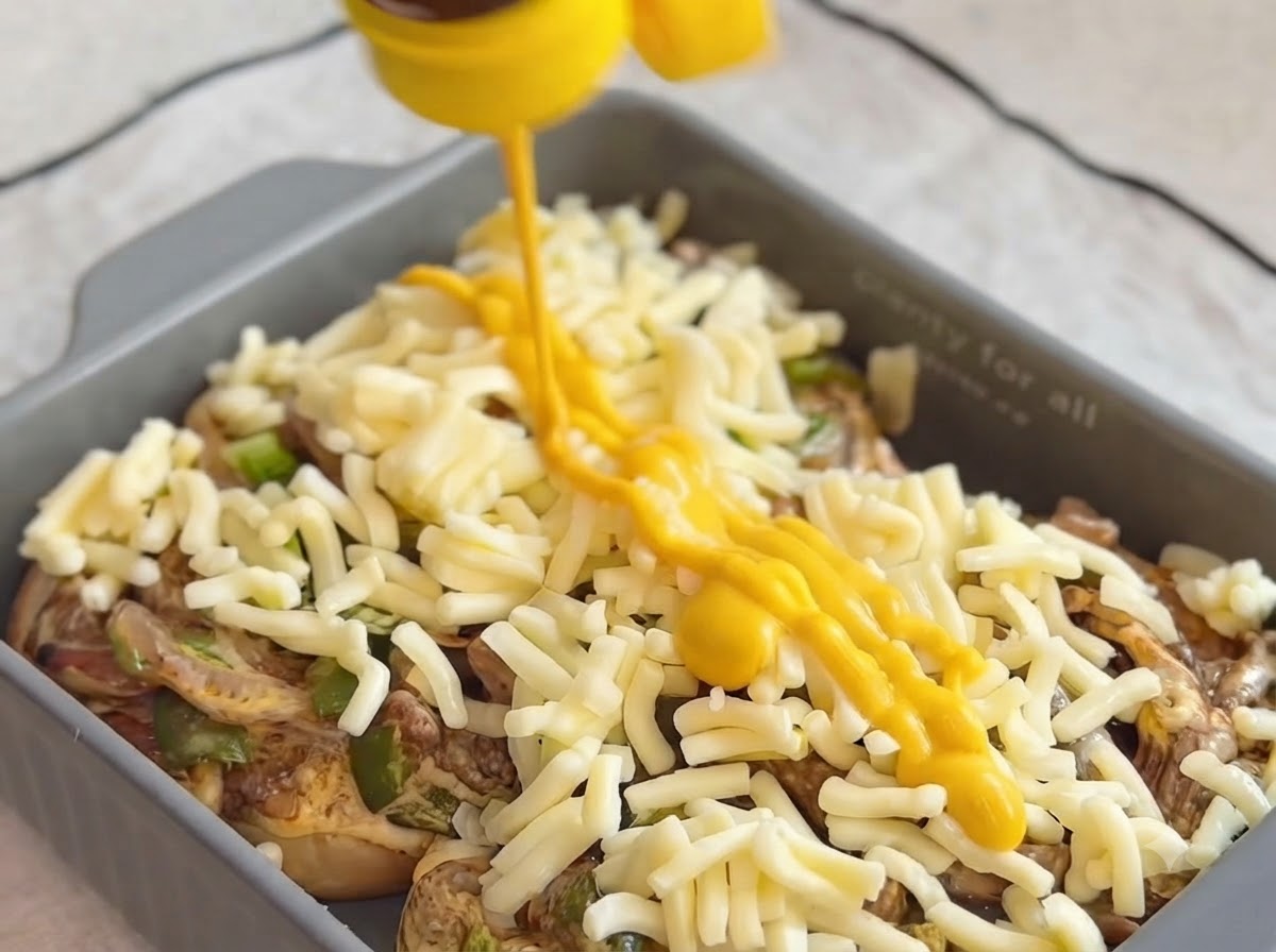 Yellow mustard sauce being squeezed in diagonal lines over the mozzarella cheese layer in a baking dish.