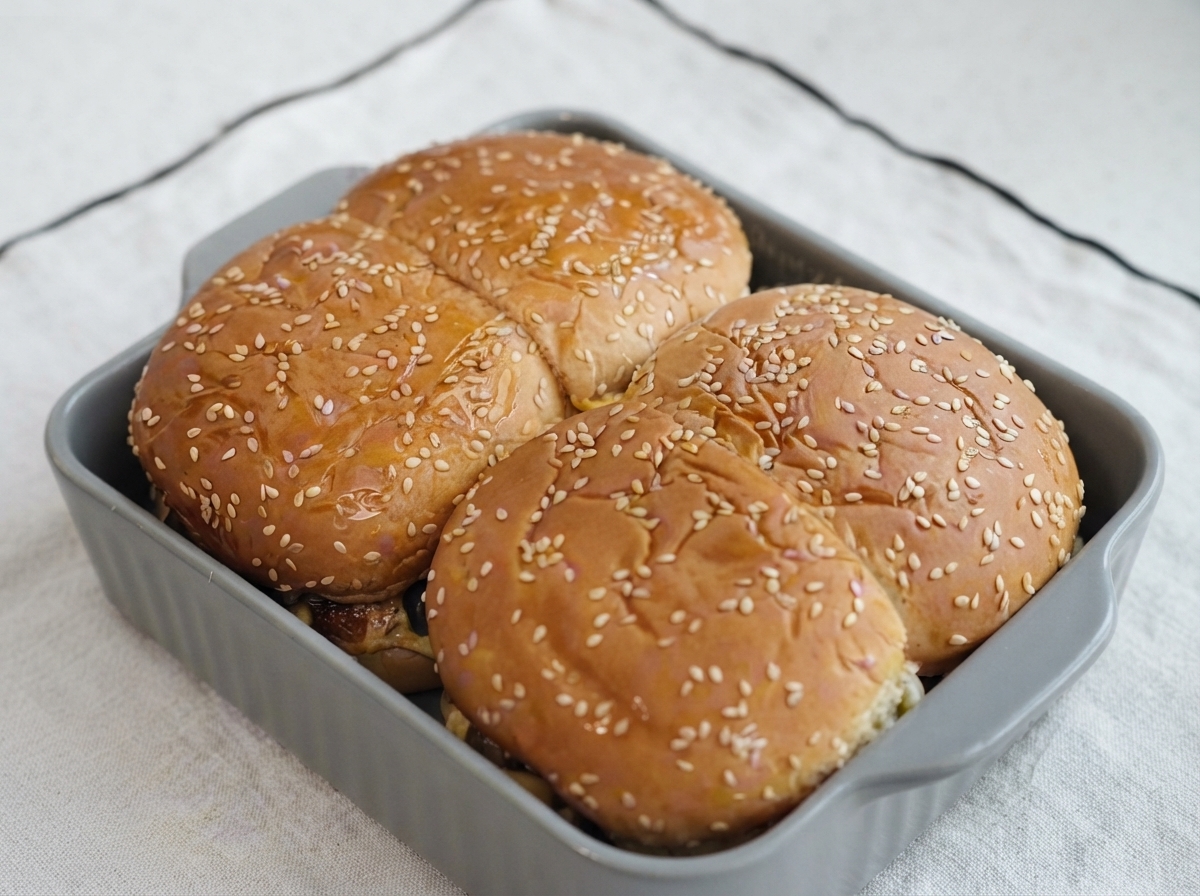 Sesame-topped burger buns inside a baking dish with oil being brushed across their surfaces.