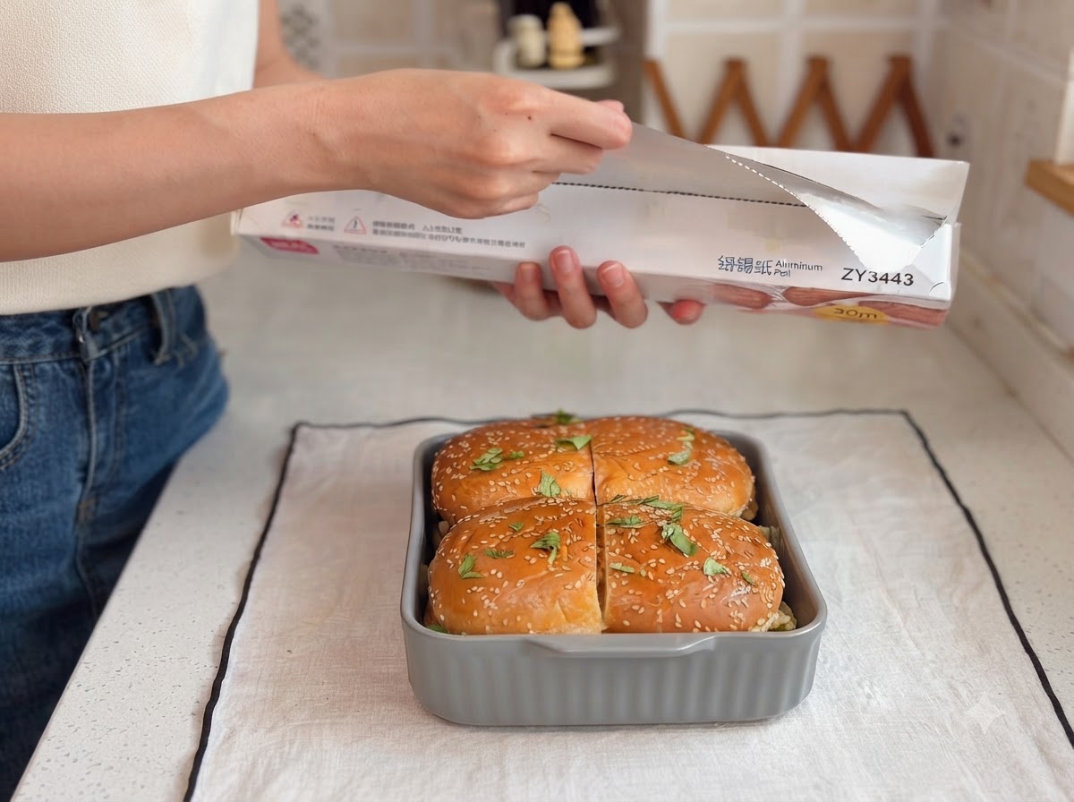 A person pulling a sheet of aluminum foil from a box above a baking dish filled with assembled burger buns.