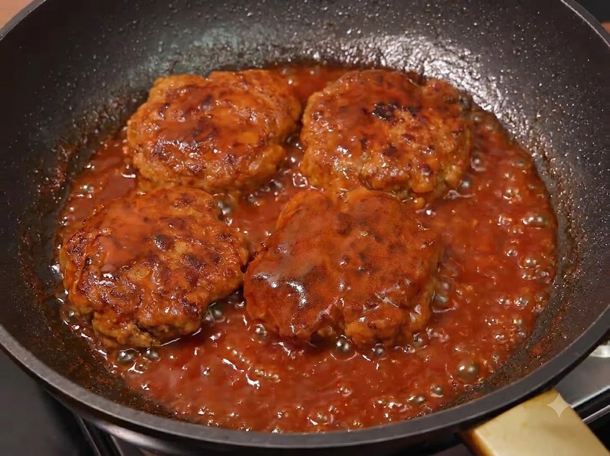 Four cooked meat patties simmering in a bubbling red sauce inside a frying pan.