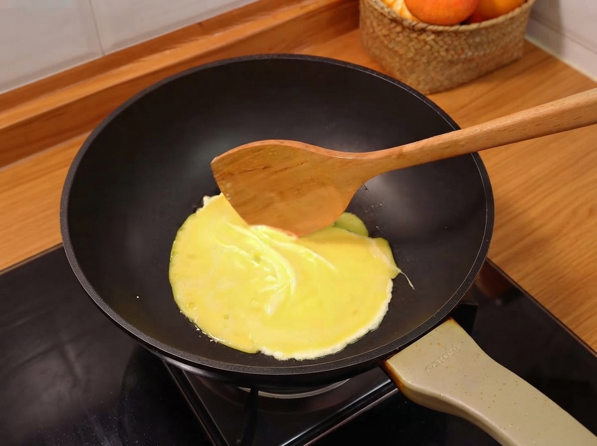 A beaten egg spreading into a thin yellow sheet being cooked in a black frying pan.
