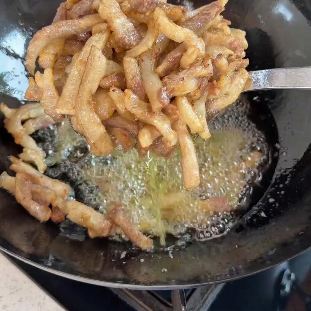 A slotted metal spoon holding up freshly fried, golden-brown pork strips over a wok of bubbling oil.