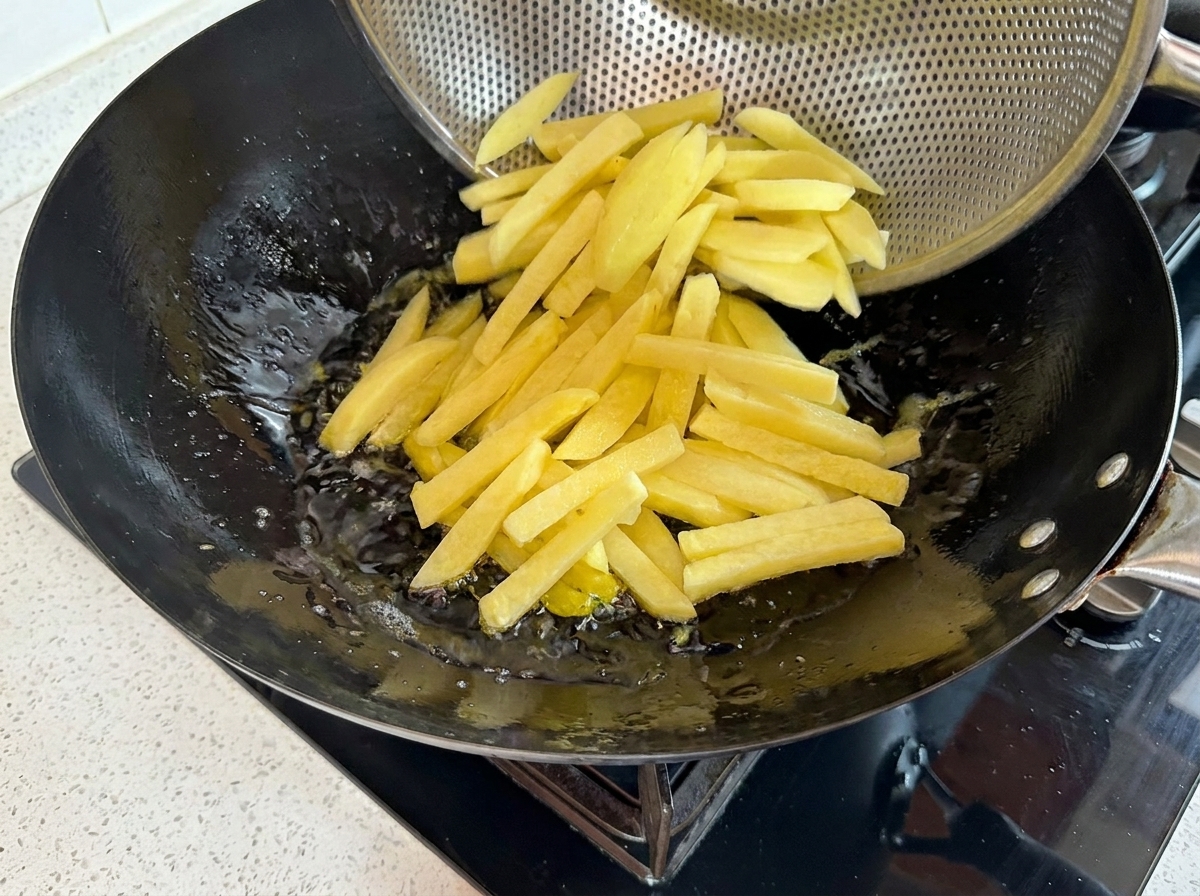 Fresh potato strips being poured from a metal strainer into a wok of hot bubbling oil.