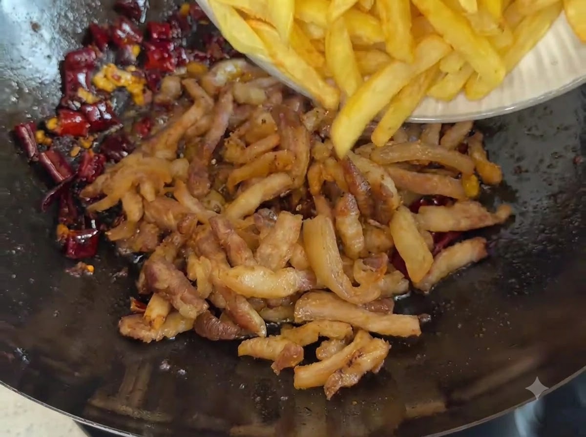 A plate sliding golden french-fry-style potato strips into a wok filled with fried pork and dark red chili spices.