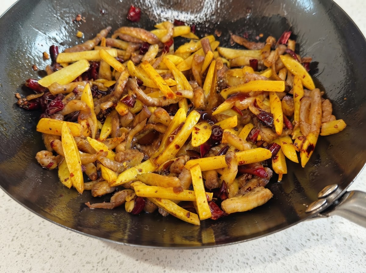 A wok filled with potato and pork strips glistening with soy sauce and sprinkled with red chili and brown spices.