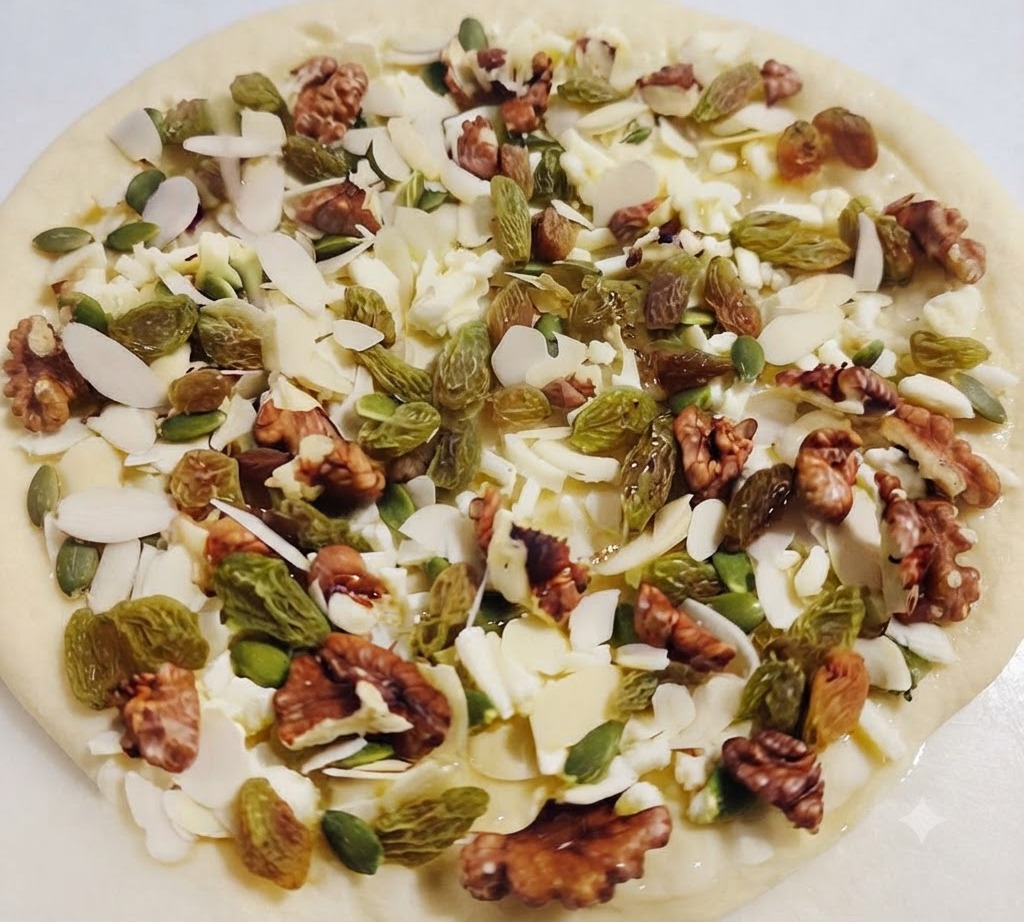 A top-down view of a pizza base sprinkled with cheese, raisins, walnuts, and almonds, with honey being drizzled over them.