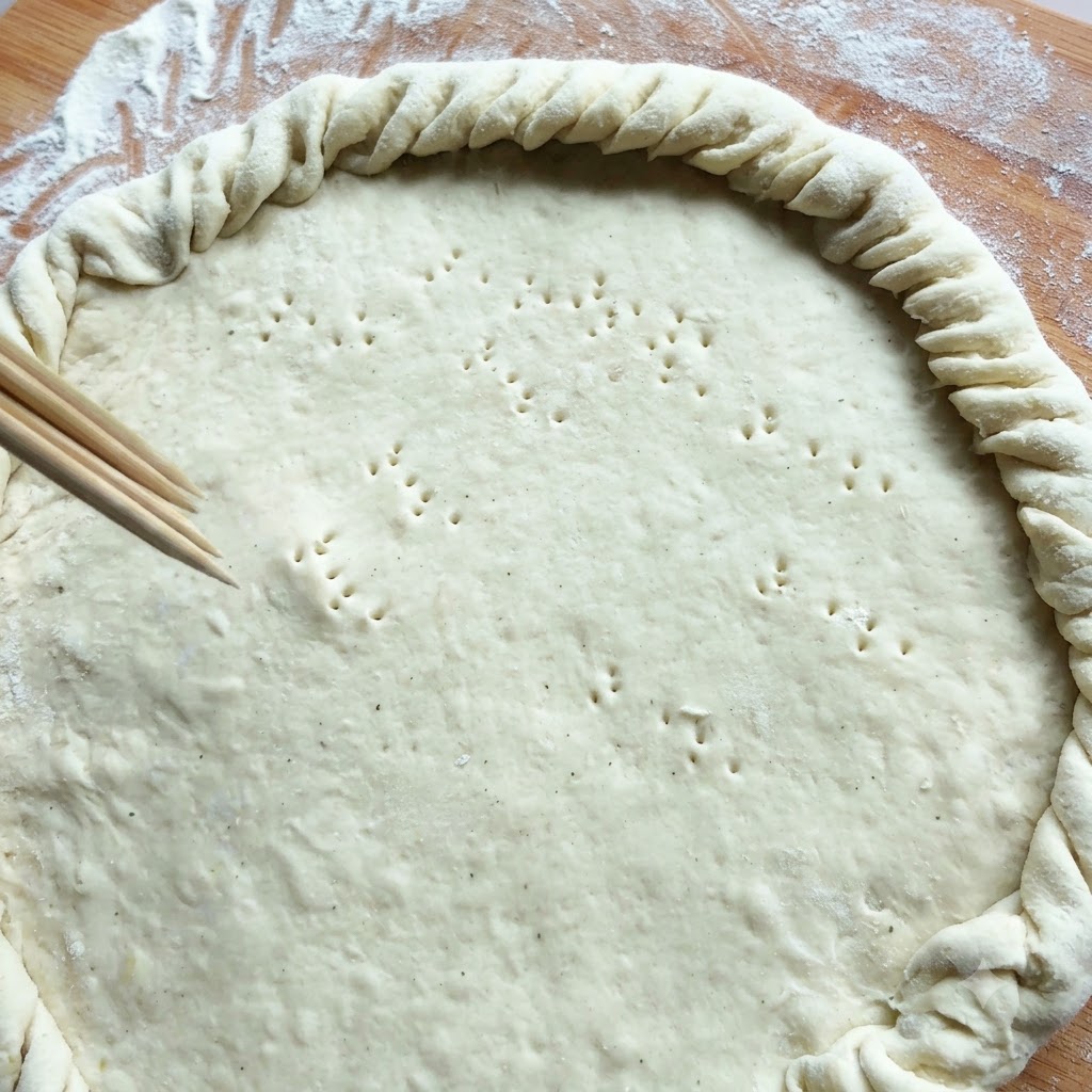 Wooden skewers poking small holes into a large circular dough crust with a twisted edge.
