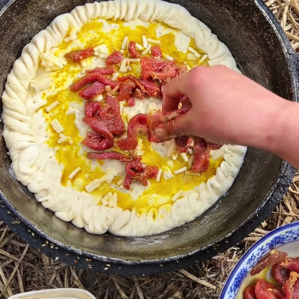 A hand placing reddish seasoned beef strips over a cheese-covered dough base in a pan.